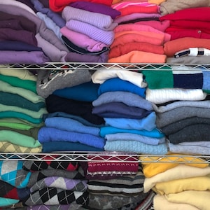 May include: A wire shelving unit filled with stacks of folded sweaters in a variety of colors including purple, pink, red, orange, green, blue, gray, yellow, and white. The garments feature various textures, solid colors, and patterns like argyle and stripes.