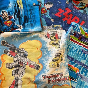 May include: A collection of fabric pieces featuring Superman in a city, Superman with the text ZAP!, a Transformers scene with Megatron and a yellow car with the text TRANSFORMERS 1984 HASBRO, and a Hot Wheels print with blue race cars.