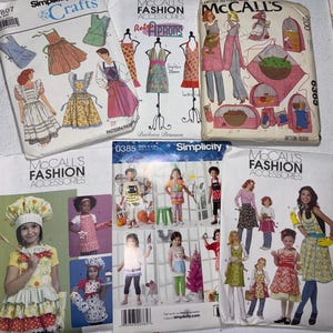 May include: A collection of six sewing pattern envelopes from Simplicity and McCall's featuring illustrations of aprons, kitchen accessories, and children's clothing. The envelopes display various designs, patterns, and instructional text.