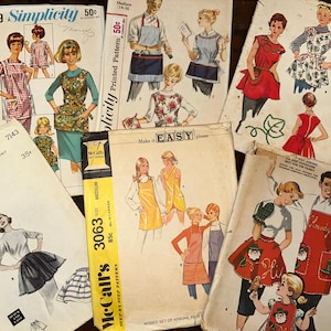 May include: A collection of vintage sewing patterns for aprons. The patterns feature different styles and designs, including a red and white gingham apron, a floral print apron, and a solid color apron. The patterns are from Simplicity and McCall's.