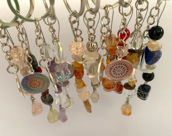 May include: A collection of colorful crystal and gemstone keychains with silver chains and charms. Each keychain features a variety of beads, including clear quartz, amethyst, rose quartz, carnelian, and agate. The charms are silver with a mandala design.