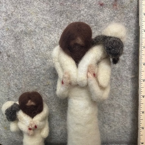 May include: Two felted wool figurines. One is a tall figure in a white robe, holding a small figure with a dark brown head and a white and gray head. The smaller figure is also in a white robe. The figures have red accents.