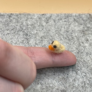 May include: A small, yellow felt duck with orange beak and black eyes. The duck is sitting on a finger.