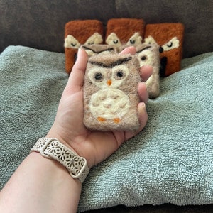 May include: A set of five felted soap bars, each featuring an owl design. The soap bars are brown and white with a fluffy texture. The soap bars are arranged in a row on a blue towel.