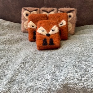 May include: A collection of handmade felt animal ornaments. The ornaments feature a fox and owl design with white, black, and brown accents. The fox ornaments are a reddish-brown color, while the owl ornaments are a light brown color.