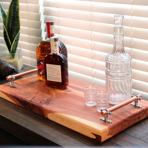 May include: A wooden serving tray with copper handles holds two bottles of liquor, a clear glass decanter, and two small glasses. The tray has a natural wood grain finish and is set against a window with blinds.