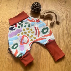 Könnte beinhalten: Graue Babyhose mit einem bunten abstrakten Muster in Rot, Blau, Grün und Lila. Die Bündchen und der Bund sind einfarbig rostfarben. Ein Tannenzapfen und ein Holzspielzeug befinden sich im Hintergrund.