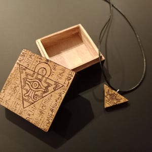 Könnte beinhalten: Holzkiste und Anhänger-Set mit graviertem Horusauge-Design. Die quadratische Box ist offen, mit passendem Deckel. Der dreieckige Anhänger hängt an einer schwarzen Kordel. Das Set ist aus hellem Holz gefertigt.