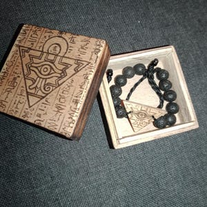 Puede incluir: Una caja de madera con un diseño tallado y una pulsera a juego con un colgante. La caja está abierta, revelando una pulsera de cuentas negras y un colgante triangular con un símbolo de ojo. La caja y el colgante son de color marrón claro.