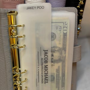 May include: A gray binder with gold hardware holds a clear plastic sleeve containing a twenty-dollar bill. The sleeve is labeled "JACOB MICHAEL ALLOWANCE & SAVINGS" and "JAKEY POO". Other binders are visible in the background.