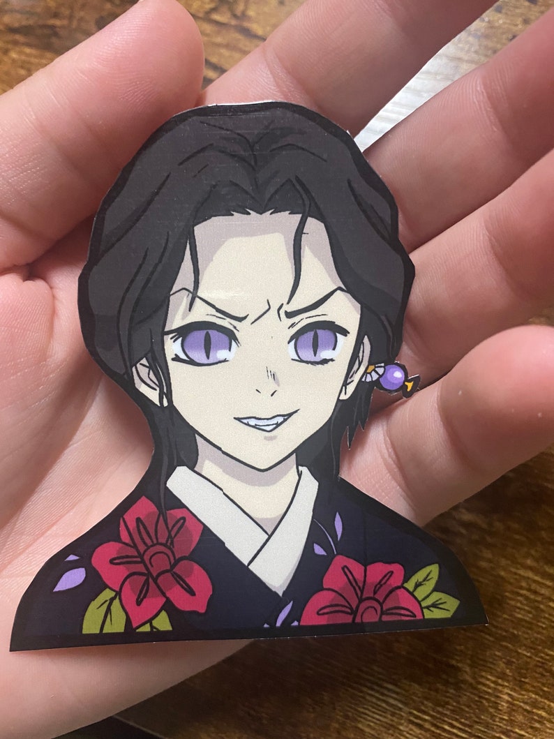 May include: A sticker of a female anime character with black hair, purple eyes, and a smirk. She is wearing a black and purple kimono with red flowers. The sticker is on a hand.