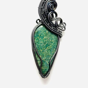 Op de afbeelding: Een groene edelsteen hanger met een zilveren draad gewikkeld ontwerp. De hanger is druppelvormig en heeft een delicaat, ingewikkeld ontwerp.