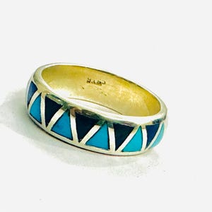 Pode incluir: Um anel de banda de prata com um padrão geométrico de pedras turquesa e pretas. O anel é marcado com "925" no interior.