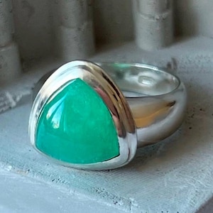 May include: A silver ring featuring a triangular green gemstone set in the centre. The ring is polished and has a smooth, rounded band.