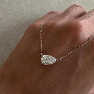 Könnte beinhalten: Eine zarte Halskette mit einem tropfenförmigen Diamantanhänger. Der klare Diamant ist an einer feinen Goldkette befestigt. Die Halskette wird vor einem neutralen Hintergrund präsentiert, um ihre Eleganz hervorzuheben.