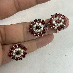 Könnte beinhalten: Ein Set aus drei blumenförmigen Ringen mit einem dunkelroten Edelstein in der Mitte, umgeben von einem Ring aus kleinen weißen Perlen und einem äußeren Rand aus dunkelroten Edelsteinen, gefasst in ein roségoldfarbenes Metallband.