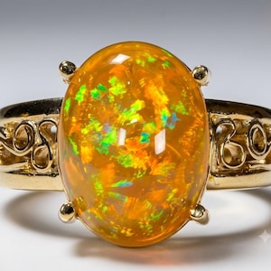 Puede incluir: Un anillo de oro con un gran cabujón de ópalo naranja de forma ovalada como pieza central. La piedra preciosa muestra destellos vibrantes de luz verde y amarilla. La banda está hecha de oro pulido e incluye intrincados patrones de volutas.
