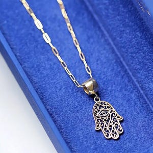 Könnte beinhalten: Goldfarbene Halskette mit einem Hamsa-Hand-Anhänger. Der Anhänger hat ein Augendesign und aufwendige filigrane Details. Die Halskette wird auf einem blauen Hintergrund präsentiert.