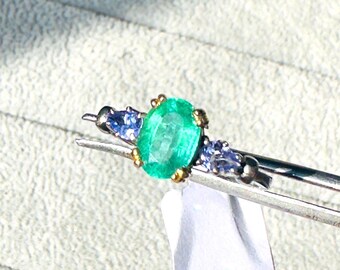 May include: A silver ring featuring an oval-cut, vibrant green emerald as the centerpiece. The emerald is flanked by two smaller, round blue gemstones. The ring has gold accents around the emerald and blue gemstones.