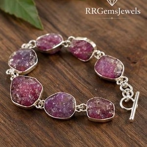 Peut inclure: Un bracelet composé de six pierres précieuses magenta brutes et irrégulières, serties dans des montures en métal argenté, reliées par des maillons circulaires argentés et fermées par un fermoir à barre. Le texte RRGemsJewels est visible.
