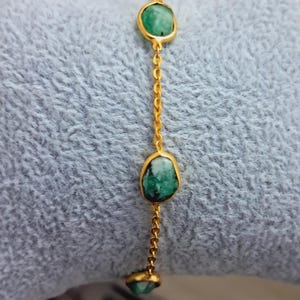 Peut inclure: Un bracelet en chaîne dorée avec trois breloques en pierres précieuses vertes. Le bracelet est sur une surface bleu clair et duveteuse.