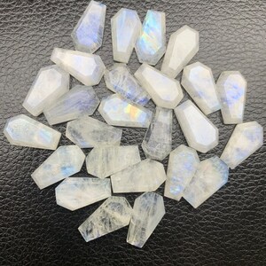 Può includere: Una collezione di 18 cabochon di pietra di luna bianca iridescente a forma di bara. Le pietre sono disposte in cerchio su una superficie nera.