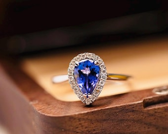 May include: A pear-shaped, blue gemstone ring with a halo of small, clear stones. The ring is set in a gold band and rests on a wooden surface. The gemstone is a deep, vibrant blue.