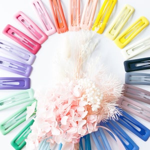 Könnte beinhalten: Eine Regenbogen-Auswahl an bunten Plastik-Haarklammern, die in einem Halbkreis auf weißem Hintergrund angeordnet sind. Die Klammern sind alle gleich groß und haben die gleiche Form, und sie sind in einem Farbverlauf von Rot nach Lila angeordnet.