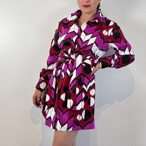 Könnte beinhalten: Ein knielanges Kleid mit geometrischem Muster in Rot, Weiß, Schwarz und Lila. Das Kleid hat lange Ärmel und eine Knopfleiste, die mit einem passenden Gürtel in der Taille zusammengebunden wird. Das Kleid wird mit schwarzen Loafern getragen.