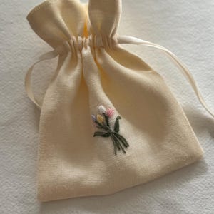 May include: A small, cream-coloured drawstring bag with a delicate embroidered floral design. The bag is made of a soft fabric and features a gathered top with a satin ribbon closure. The embroidery includes pink, yellow, and purple flowers.