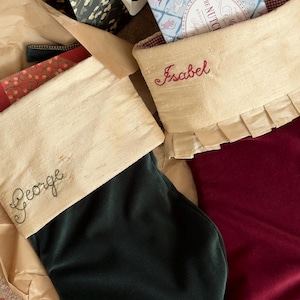 May include: Two velvet Christmas stockings with cream-colored cuffs. One stocking is dark green with the name George embroidered in brown, and the other is deep red with the name Isabel embroidered in red and a ruffled trim along the cuff.