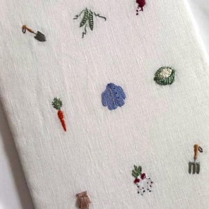 May include: A white fabric panel with various embroidered images of garden vegetables and tools, including a carrot, peas, a shovel, a rake, a beetroot, and a cabbage.