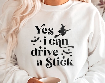May include: White sweatshirt with the text "Yes I can drive a Stick" in black, along with witch and bat graphics. The sweatshirt is a Halloween-themed item.