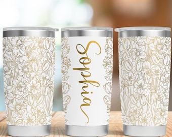 May include: Three white insulated tumblers with silver lids. Two tumblers feature a gold floral pattern, while the center tumbler has the name "Sophia" in gold script. The tumblers are on a wooden surface.