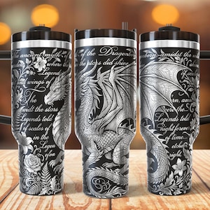 Op de afbeelding: Een zwarte roestvrijstalen beker met handvat, voorzien van een gedetailleerde witte gegraveerde draak, bloemmotieven en cursieve tekst met de woorden The ancient myth of the Dragonborn, where the stars did shine, and legends told.