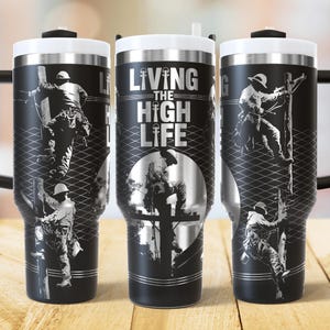 Könnte beinhalten: Schwarzer isolierter Becher mit weißem Deckel und Griff. Der Becher zeigt eine Grafik eines Elektrikers, der einen Strommast erklimmt, mit den Worten "LIVING THE HIGH LIFE" in silberner Schrift.