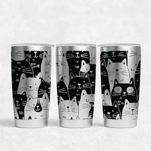 Könnte beinhalten: Drei Edelstahl-Reisebecher mit einem schwarz-weißen Katzenmuster. Das Design zeigt ein sich wiederholendes Muster stilisierter Katzenköpfe. Die Katzen sind in Schwarz und Weiß gehalten, wodurch ein kontrastreiches Design entsteht. Die Becher haben einen silbernen Rand und Boden.