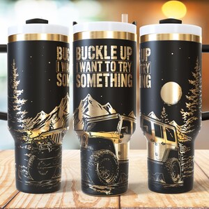 May include: Black stainless steel travel mug with a gold Jeep design, mountains, and trees. The mug has the text "BUCKLE UP I WANT TO TRY SOMETHING". It has a white lid with a gold rim and a black handle.