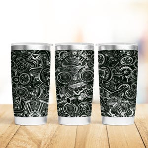 May include: Three stainless steel tumblers with a black and white graphic print of a car engine. The tumblers are on a wooden surface.