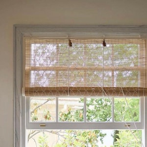 Könnte beinhalten: Ein weißes Fenster mit einem braunen Bambusrollo mit geometrischem Muster. Das Rollo ist hochgezogen und gibt den Blick auf grüne Bäume vor dem Fenster frei.