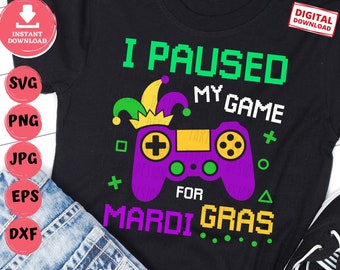 Puede incluir: Camiseta negra con un gráfico de mando de videojuego morado y verde. El texto "I Paused My Game For Mardi Gras" está escrito en letras pixeladas verdes y blancas.