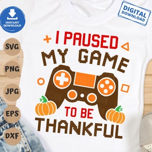 Op de afbeelding: Een wit T-shirt met een bruine videogamecontroller en de tekst "Ik heb mijn game gepauzeerd om dankbaar te zijn" met twee oranje pompoenen aan weerszijden van de tekst.
