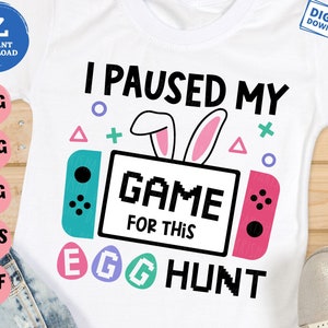 May include: White t-shirt with a graphic design of a video game controller with bunny ears and the text "I paused my game for this egg hunt".