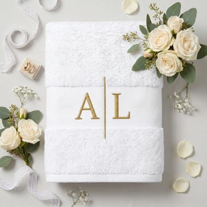 Könnte beinhalten: Weißes Badetuch mit gestickten goldenen Initialen "AL". Das Handtuch ist von weißen Rosen, Grün und dekorativen Bändern umgeben. Das Handtuch ist aus einem weichen, flauschigen Material, perfekt als Hochzeits- oder Einzugsgeschenk.
