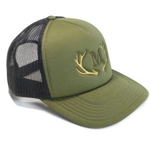 Puede incluir: Gorra de camionero verde oliva con parte trasera de malla negra. La parte delantera presenta un diseño dorado bordado de cuernos y la letra "M". La gorra tiene una visera curva y un botón en la parte superior.