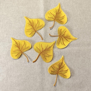 Könnte beinhalten: Sechs gelbe, blattförmige, gestickte Aufnäher mit braunen Adern und Stielen sind auf einem hellgrauen Stoff angeordnet. Die Blätter haben eine herzähnliche Form, und die Stickerei zeigt die Adern.