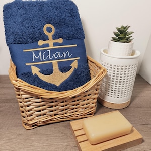 Può includere: Un asciugamano blu navy con un motivo di ancora dorata e il nome "Milan" ricamato, presentato in un cestino di vimini. Sono visibili anche una piccola pianta succulenta in un vaso bianco e una saponetta su un portasapone di legno.