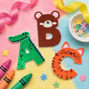 Peut inclure: Trois écussons en tissu brodé en forme de lettres A, B et C. La lettre A est un alligator vert, la lettre B est un ours brun et la lettre C est un tigre orange. Les écussons présentent des coutures détaillées et des traits d'animaux.