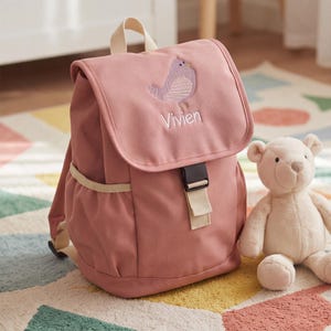 Könnte beinhalten: Ein altrosa Rucksack mit einer oberen Klappe, die einen gestickten lila Vogel und den in weißem Garn gestickten Namen Vivien zeigt. Der Rucksack verfügt über einen beigen Tragegriff, beige Seitentaschen und einen schwarzen Kunststoffschnallenverschluss.