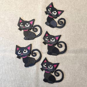 Op de afbeelding: Vijf zwarte kattenpatches met roze accenten, waaronder oren en kragen, zijn gerangschikt op een neutrale stoffen achtergrond. Elke kat heeft een gestileerd ontwerp met een gekrulde staart en witte snorharen. Geschikt voor knutselprojecten.